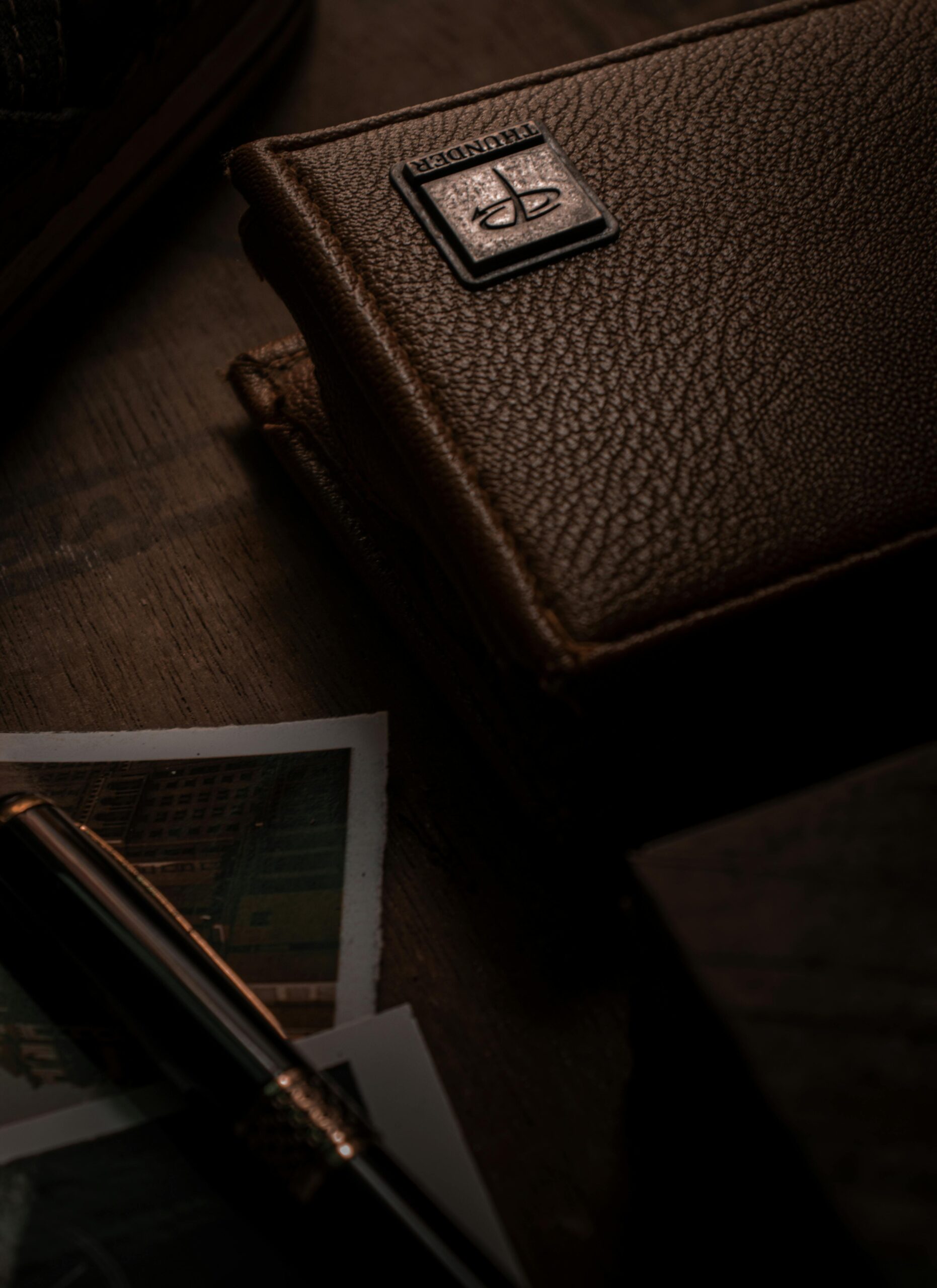 Close-up of a textured brown leather wallet on a wooden desk beside a pen and photos.